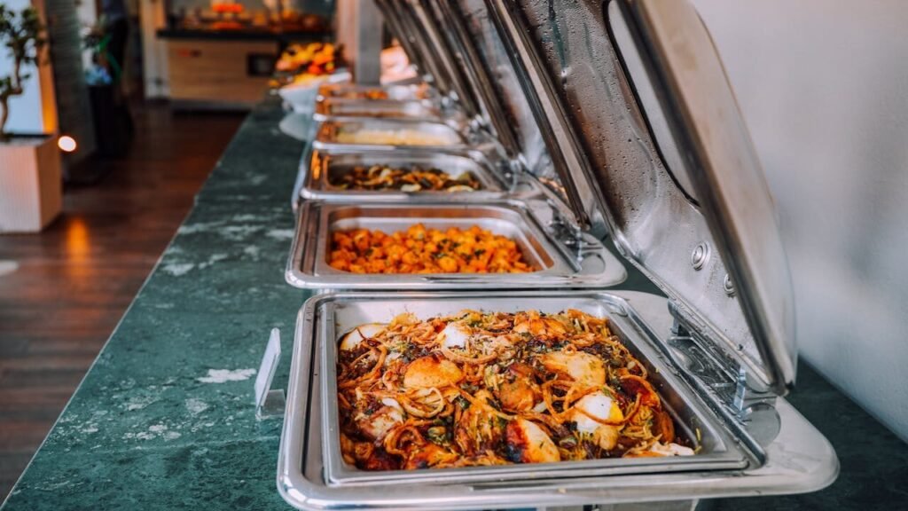 Flaming Kabob A buffet setup with stainless steel chafing dishes containing various cooked dishes such as pasta, vegetables, and other mixed cuisines. The setting appears to be indoors with a focus on the food. Commerce TWP, MI